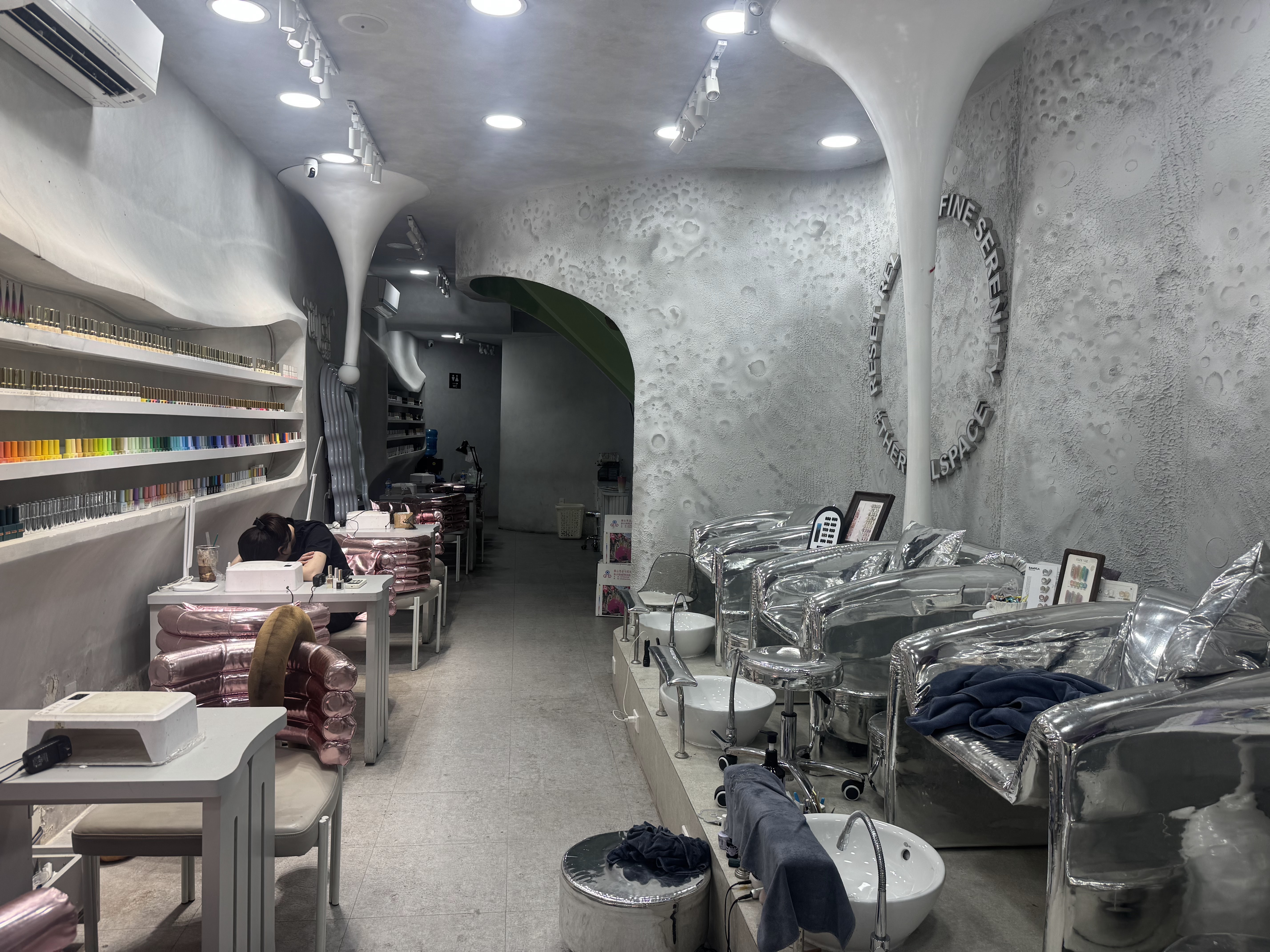Quầy nail bằng gỗ tối, sơn gel xếp gọn theo dải màu, đèn rọi tập trung lên mặt bàn.